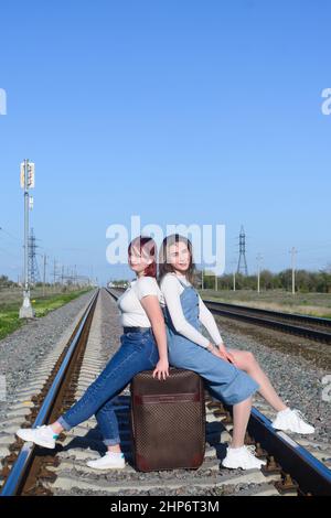 Two young women are sitting on large suitcase. Railroad rails on background of blue sky. Travel concept. Stock Photo