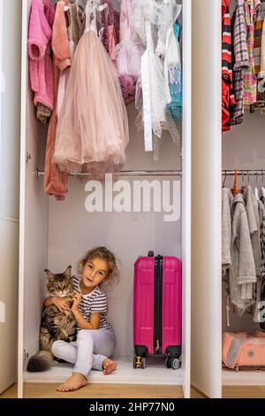 Funny little girl hiding in cupboard hugging cat feeling love and positive emotion Stock Photo