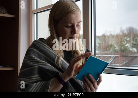 Woman reading a bright blue book at the window in winter dressed in woolen scarf Stock Photo
