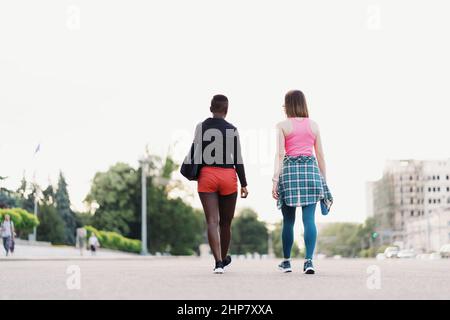 Friends back view in sportswear walking after a sport session in the city discussing . Multiethnic women having a fitness workout jogging. Stock Photo