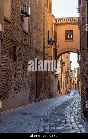 Old Street - Evening view of an old narrow winding cobblestone street, with a brick bridge at above, in Jewish Quarter of historic city Toledo, Spain. Stock Photo