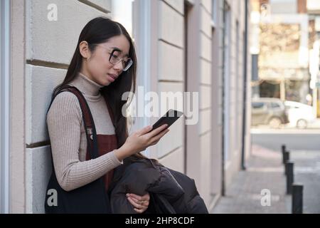Portrait of a young asian woman using the phone standing up against the wall. Stock Photo