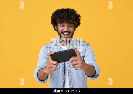 Excited young indian man using smartphone playing game on yellow background. Stock Photo
