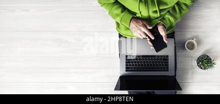 Businessman in a green hoodie with laptop and smartphone works while sitting on a wooden floor. Copy space for text. Top view. Stock Photo