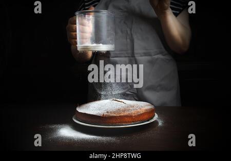 Woman wearing apron sprinkle with powdered sugar sweet cake. House wife making homemade pie. Stock Photo