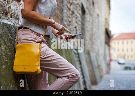 Fashionable woman is leaning against wall and using a smartphone. Stylish woman wearing trousers and t-shirt and holding a smart phone. Stock Photo