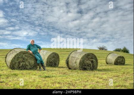 Farmer makes after hay harvest a break and enjoy the moment between the round hay bales on the mown meadow. Stock Photo