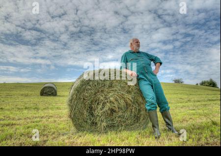 Farmer makes a break and leans against a round hay baleand on the mown meadow. Stock Photo