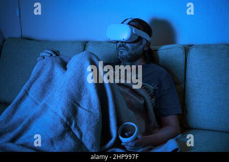 Man sitting on sofa uses virtual reality headset at night, sad person looking in VR glasses at home alone. Concept of loneliness, watching tv, relaxat Stock Photo