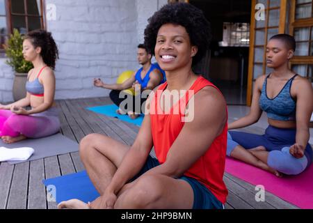 Portrait of smiling young multiracial man sitting against friends practicing yoga in backyard Stock Photo