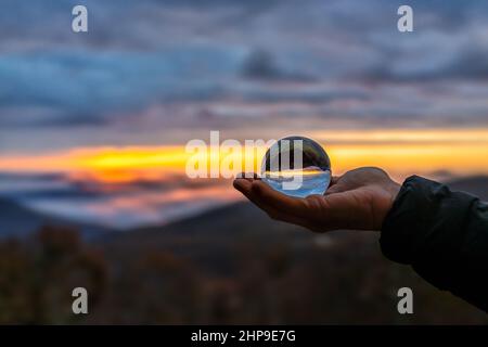 Hand holding crystal glass lens ball in palm view of round globe with reflection of fog mist clouds over blue ridge mountains at colorful sunrise in W Stock Photo