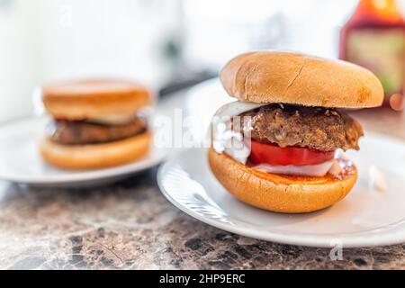 Two small burgers closeup of one with hamburger patty, buns onions and sliced tomatoes on white plates on kitchen bar counter for serving Stock Photo