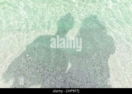 Naples beach in southwest Florida with turquoise glass green water on sunny day with shadow silhouette of two romantic people couple on sand seashells Stock Photo
