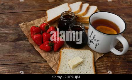 White cup of black tea with lemon. Sandwiches or toasts with butter. Strawberry and jam jar on wooden table. Breakfast and happy morning concept Stock Photo