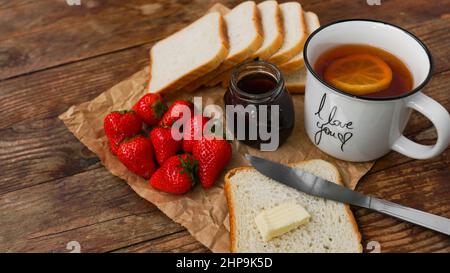 White cup of black tea with lemon. Sandwiches or toasts with butter. Strawberry and jam jar on wooden table. Breakfast and happy morning concept Stock Photo
