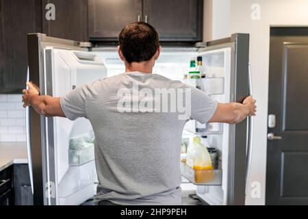 Back of hungry man opening fridge refrigerator doors domestic appliance searching for food inside with condiments and juice in modern kitchen Stock Photo