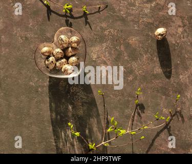 Spring Easter background with fresh leaves on twigs. Quail eggs in heart glass bowl on dark brown. Stock Photo