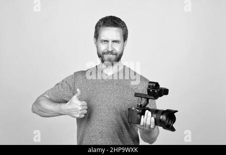 cheerful bearded man videographer making movie for vlog with camcorder showing thumb up, filming Stock Photo