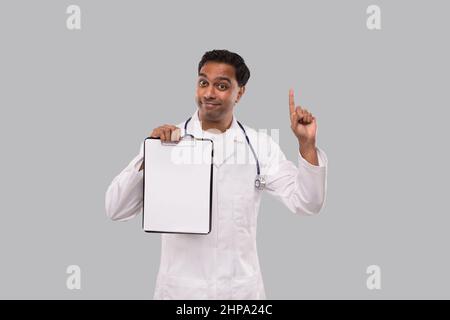 Doctor Pointing Up Clipboard Isolated. Indian Man Doctor Blank Clipboard in Hands. Male Doctor with Great Idea Stock Photo