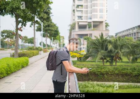 Young caucasian man in casual clothes with the backpack standing outdoors and looking at his phone. Using smartphone in the park. High quality photo Stock Photo