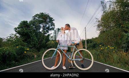 Biking road trip. Love couple on blue bike in white clothes on forest road. Just married woman and man kiss, hugs, stand on bicycle. Wedding, honeymoon. Cycling Cycle Fix. Asia Thailand ride tourism. Stock Photo