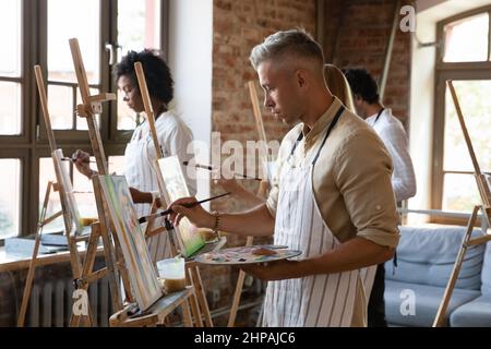 Concentrated young handsome man drawing on easel in studio. Stock Photo