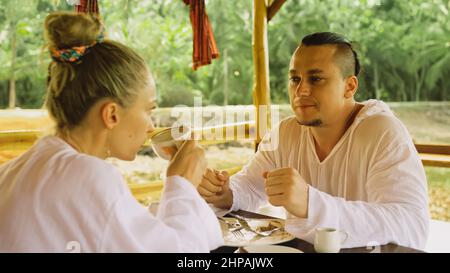 Attractive young man and woman eat and drink tasty beverages spending time in traditional local floating cafe on water. Loving happy couple having breakfast outdoors, feed each other. Tropical view Stock Photo