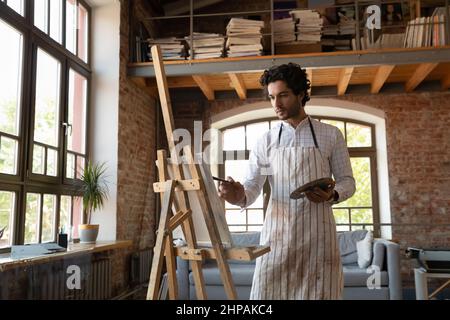 Concentrated inspired young curly man painting on canvas. Stock Photo