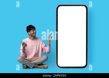 Happy indian guy in casual sitting on floor and pointing at huge smartphone with white blank screen, using mobile phone, blue studio background, full Stock Photo