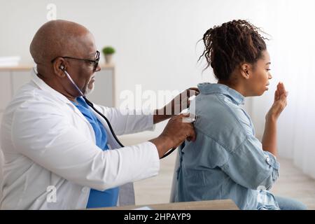 Pneumology Consultation Concept. Black male general practitioner examining coughing female patient in modern clinic, using stethoscope, listening lung Stock Photo