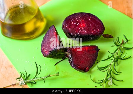 Three pieces of baked betroot, rosemary twig and glass of olive oil on green kitchen board on table. Stock Photo