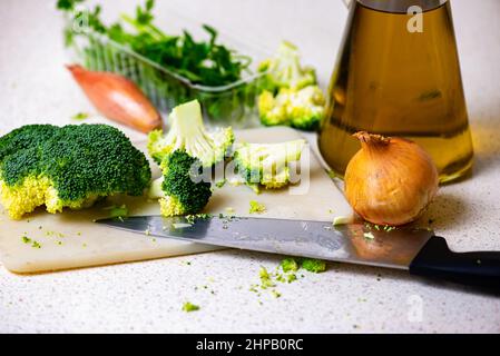 Broccoli florets, onions, parsley, bottle with olive oil and knife on kitchen board on table. Stock Photo
