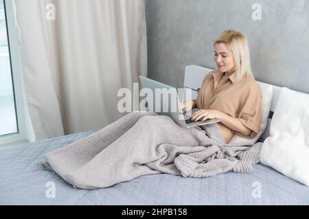 Sick ill young woman feel cold covered with blanket sit on sofa watching movie on laptop, annoyed girl shiver freezing warming at home wrapped with Stock Photo