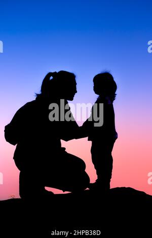 Silhouette side view of young mother and child hikers enjoying the view at the top of a mountain. Colorful sunset sky background. Friendly family. Stock Photo