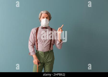 Senior man in medical mask with walking stick pointing at something on grey background Stock Photo