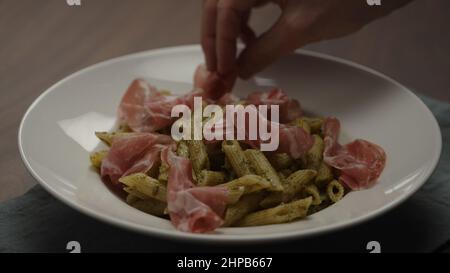 man add coppa to pesto penne in white plate, wide photo Stock Photo