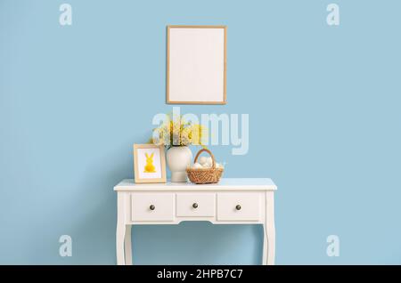 Basket with Easter eggs, mimosa flowers on table and blank photo frame near color wall Stock Photo