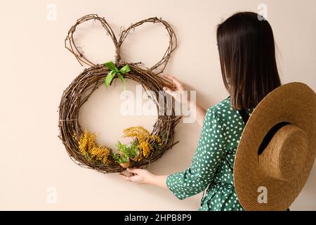 Woman hanging beautiful Easter wreath with mimosa flowers on color wall Stock Photo