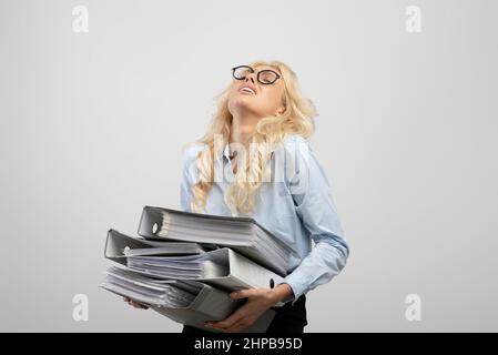 Tired female office worker holding folders of documents, overloaded with paperwork, light grey studio background Stock Photo
