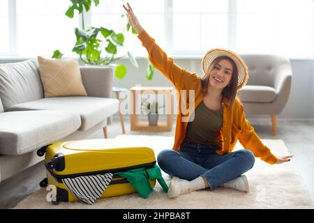 Cheery young lady sitting on floor near packed suitcase, making airplane gesture, feeling happy about abroad vacation at home, copy space. Traveling o Stock Photo