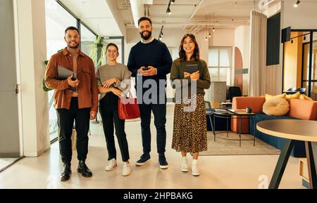Four young businesspeople smiling at the camera while standing together in an office lobby. Group of multiethnic entrepreneurs working as a team in a Stock Photo