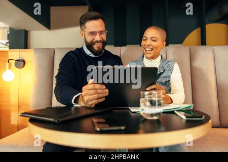 Two cheerful entrepreneurs taking a video call on a digital tablet. Two young businesspeople smiling happily during an online conference with their as Stock Photo