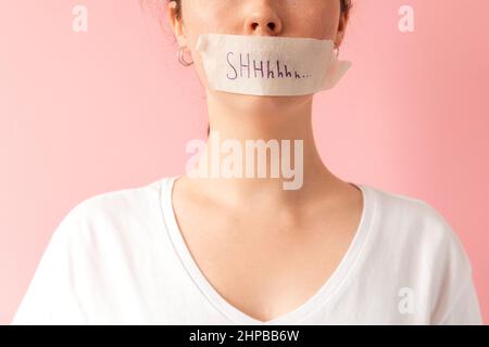 Close up of caucasian woman's face with her mouth taped shut. Tape has the inscription Shhh. Pink background. The concept of women's secrets and probl Stock Photo