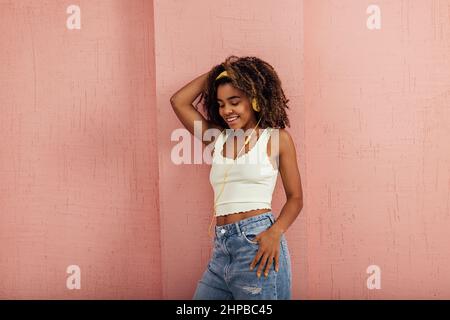 Cheerful woman with curly hair leaning pink wall while listening music Stock Photo