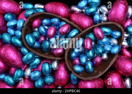 Pile of different sizes of colourful foil wrapped chocolate easter eggs in pink, red, silver and blue with two halves of a large chocolate egg. Stock Photo