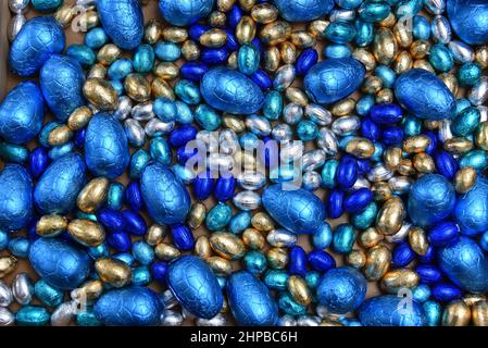 Small pale and dark blue, silver and gold foil wrapped chocolate easter eggs, with large blue eggs against a pale wood background. Stock Photo