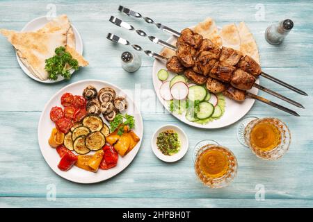 Three servings of appetizing homemade shish kebab, sliced cucumbers and radishes, barbecue vegetables, glasses of wine top view on a blue wooden backg Stock Photo