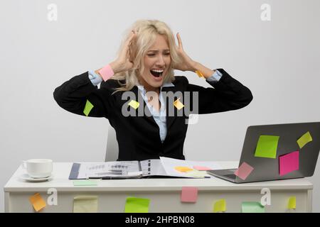 Irritated female office worker covered in sticky papers, got trouble deadline, sitting at workplace looking at laptop and touching head, light grey st Stock Photo