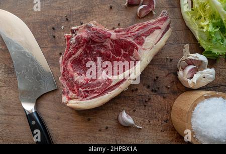 Delicious dry aged raw rubia gallega beef steak ready-to-be-cooked on a wooden board with knife, sea salt flakes, pepper, garlic, green salad Stock Photo