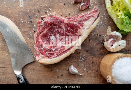 Delicious dry aged raw rubia gallega beef steak on a wooden board with knife, green salad and seasoned with sea salt flakes, pepper, garlic Stock Photo